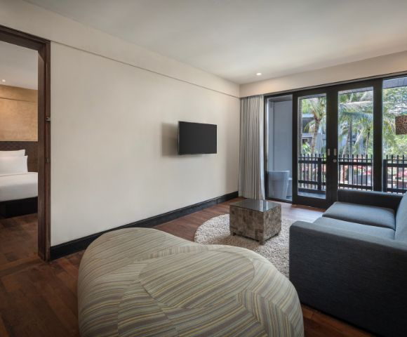Modern hotel suite with wooden floors, featuring a cozy living area with a gray sofa, TV, and pouf. Bedroom with a plush bed is visible; balcony with palm tree view.