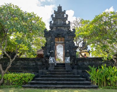 A traditional Balinese black stone gate adorned with intricate carvings and flanked by leafy trees under a blue sky, conveying a serene ambiance.