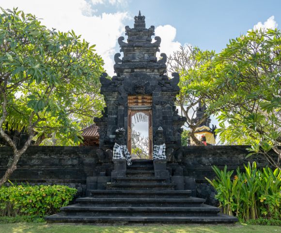A traditional Balinese black stone gate adorned with intricate carvings and flanked by leafy trees under a blue sky, conveying a serene ambiance.