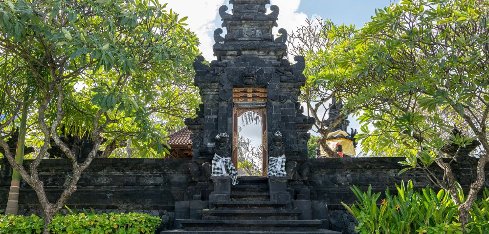A traditional Balinese black stone gate adorned with intricate carvings and flanked by leafy trees under a blue sky, conveying a serene ambiance.
