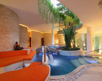Luxurious indoor spa with stone walls, orange lounge chairs, and a serene pool surrounded by lush greenery. Warm lighting creates a tranquil ambiance.