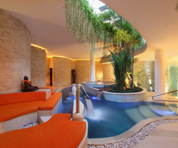 Luxurious indoor spa with stone walls, orange lounge chairs, and a serene pool surrounded by lush greenery. Warm lighting creates a tranquil ambiance.