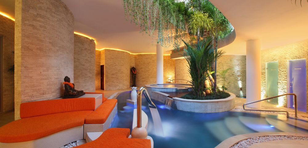 Luxurious indoor spa with stone walls, orange lounge chairs, and a serene pool surrounded by lush greenery. Warm lighting creates a tranquil ambiance.
