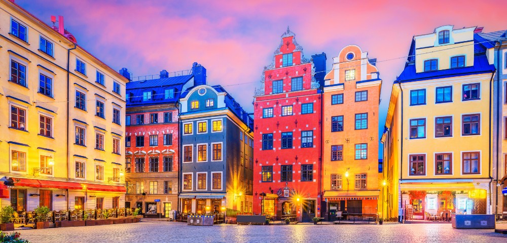 Colorful historic buildings line a cobblestone street under a vibrant sunset sky in Stockholm's Gamla Stan, creating a lively and charming atmosphere.