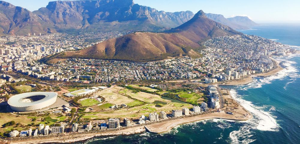 Aerial view of Cape Town with Table Mountain in the background, the iconic stadium, and coastline. Urban landscape meets lush mountains and ocean.