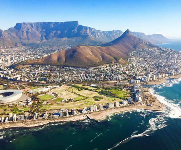Aerial view of Cape Town with Table Mountain in the background, the iconic stadium, and coastline. Urban landscape meets lush mountains and ocean.
