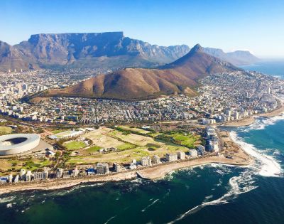 Aerial view of Cape Town with Table Mountain and Lion's Head in the background, cityscape and coastline in the foreground, under a clear blue sky.