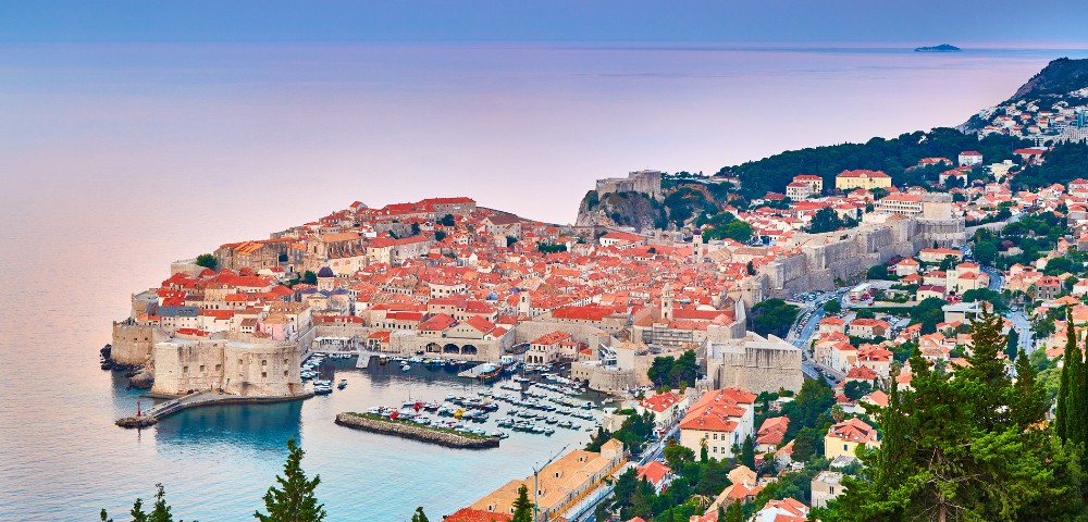 Scenic view of Dubrovnik, Croatia at sunset with terracotta rooftops, ancient stone walls, and a calm sea, conveying a historic and tranquil atmosphere.