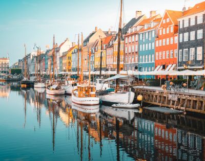 Colorful buildings line a canal in Copenhagen. Boats are docked along the water, reflecting vibrant façades under a clear blue sky, creating a lively, charming scene.