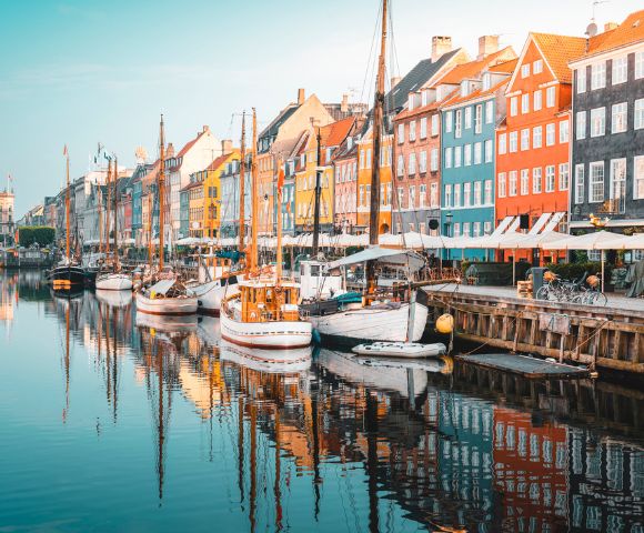Colorful buildings line a canal in Copenhagen. Boats are docked along the water, reflecting vibrant façades under a clear blue sky, creating a lively, charming scene.