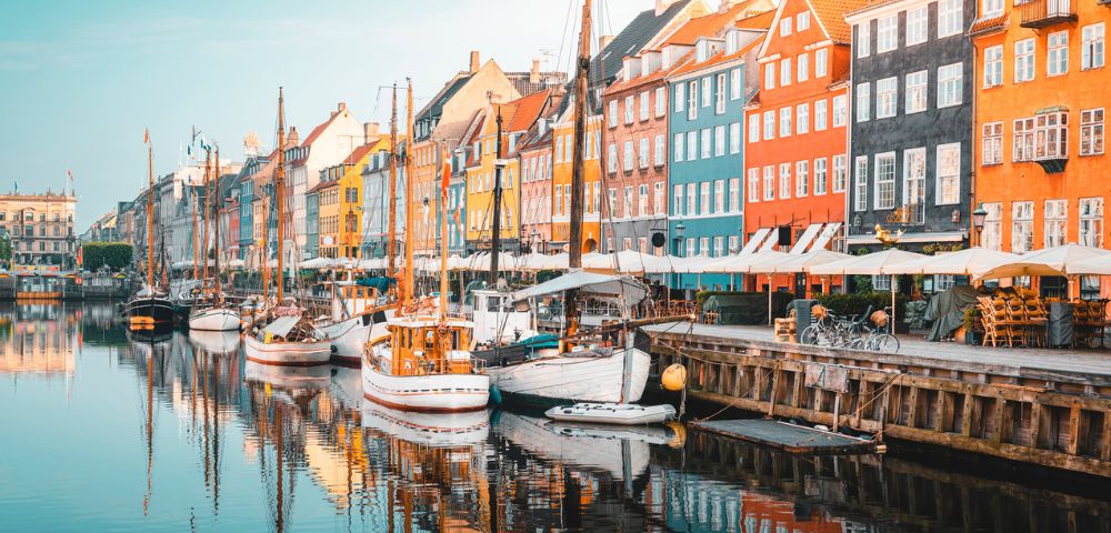 Colorful buildings line a canal in Copenhagen. Boats are docked along the water, reflecting vibrant façades under a clear blue sky, creating a lively, charming scene.