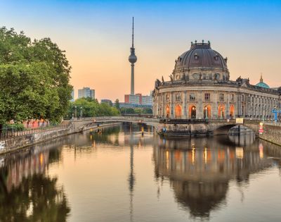 Sunset view of Berlin's Museum Island with the Bode Museum and TV Tower, reflecting in the tranquil river, framed by lush green trees.