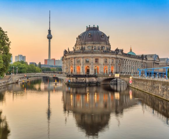 Sunset view of Berlin's Museum Island with the Bode Museum and TV Tower, reflecting in the tranquil river, framed by lush green trees.
