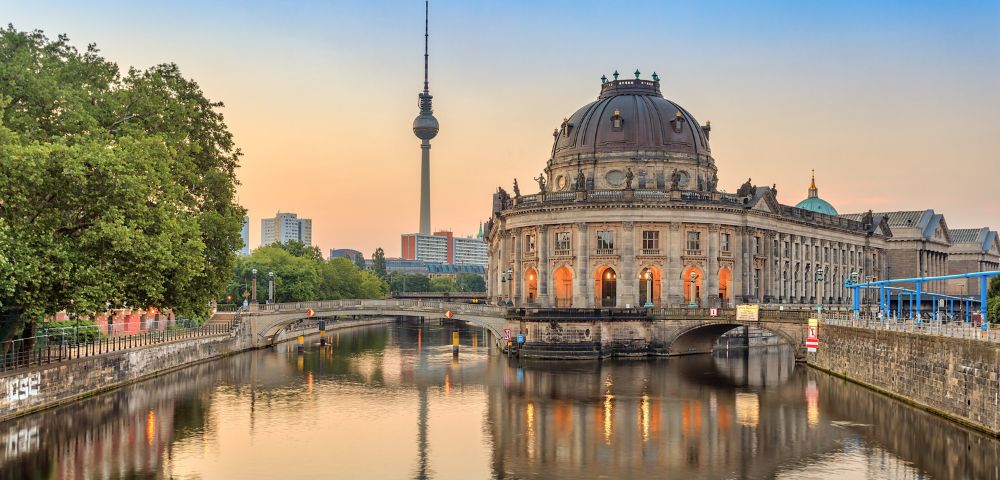 Sunset view of Berlin's Museum Island with the Bode Museum and TV Tower, reflecting in the tranquil river, framed by lush green trees.