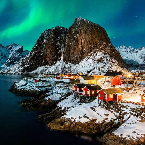 Snow-covered village with red cabins by a fjord under a vivid green aurora borealis. Majestic mountains surround, creating a serene, magical atmosphere.