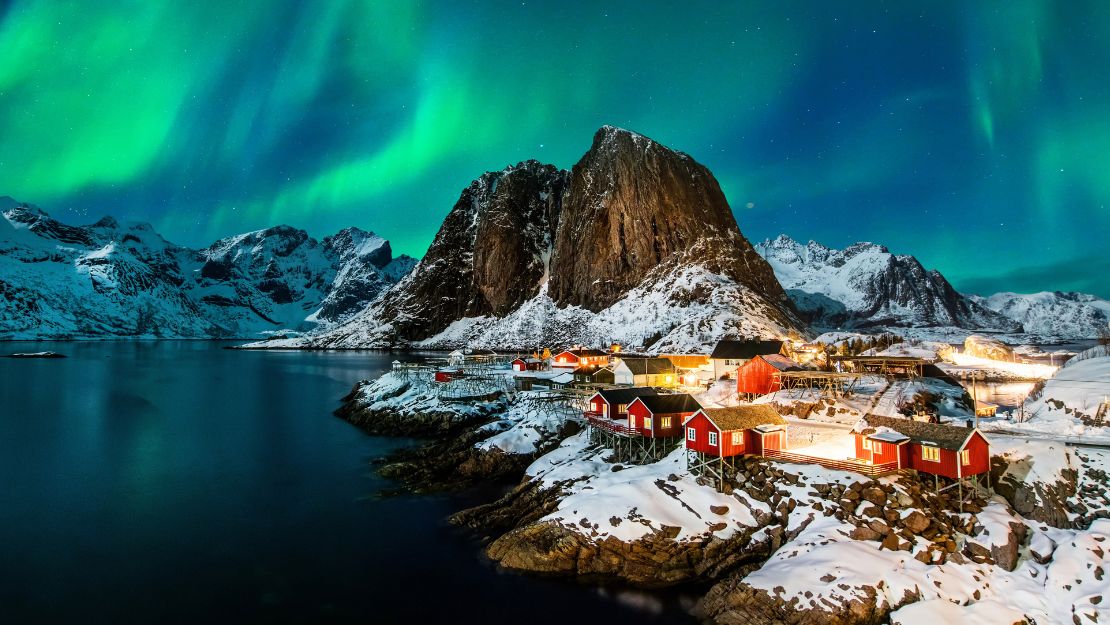 Snow-covered village with red cabins by a fjord under a vivid green aurora borealis. Majestic mountains surround, creating a serene, magical atmosphere.