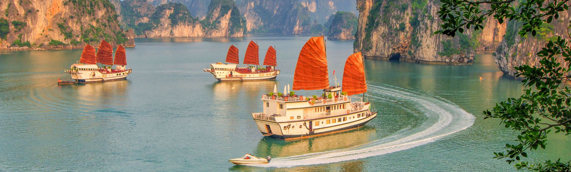 Traditional boats with striking red sails navigate calm, turquoise waters surrounded by towering limestone cliffs. A small speedboat trails nearby, under a sky at golden hour.