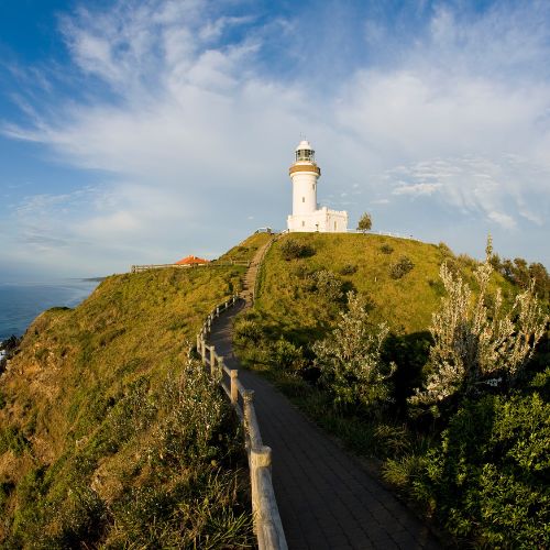 A solitary white lighthouse stands atop a grassy cliff, overlooking a vast, calm ocean under a serene blue sky. A winding path leads to it.