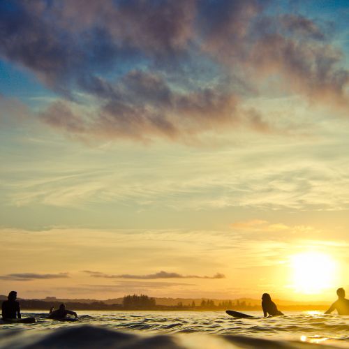 Surfers sit on their boards in calm ocean water under a vibrant sunset sky. Clouds are softly painted in hues of orange and blue, conveying a peaceful mood.