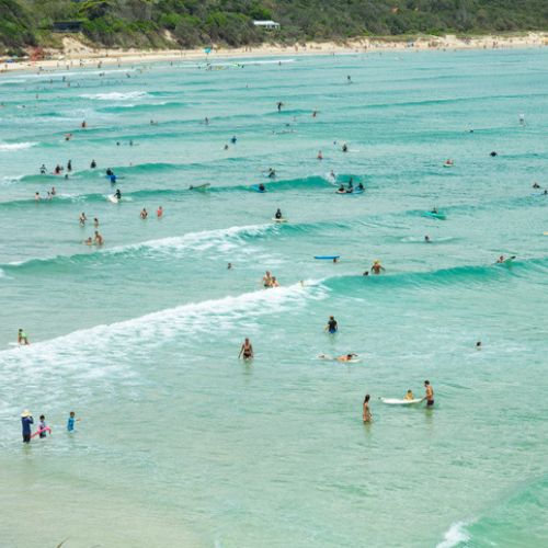 Beach scene with many people enjoying the ocean. Soft waves and turquoise water create a lively, tranquil atmosphere. Distant sandy shore with scattered swimmers.