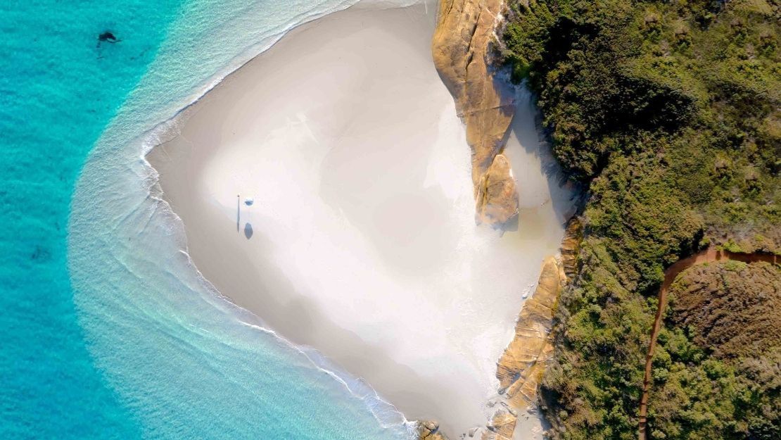 Aerial view of a secluded, heart-shaped beach with white sand and clear turquoise water, bordered by lush greenery and rocky cliffs, creating a serene and idyllic atmosphere.