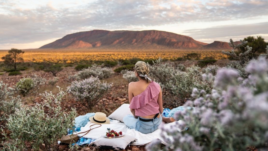 A woman in a pink, one-shoulder top sits on a picnic blanket in a vast desert landscape. She overlooks a mountain at sunset, evoking a serene and contemplative mood.