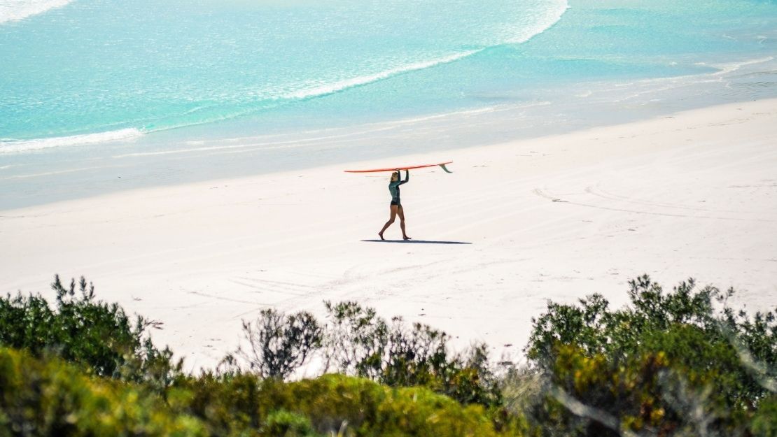 Surfer carrying a surfboard on a wide, empty sandy beach with turquoise ocean waves in the background.