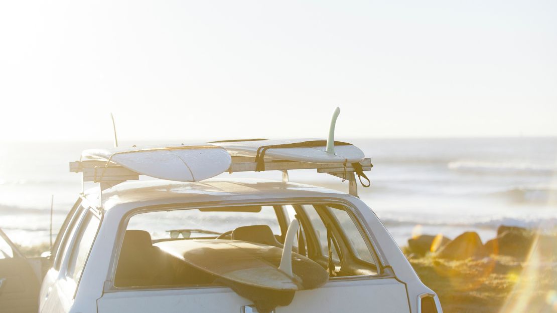 White car parked by the beach with surfboards on its roof rack and inside, ocean waves and rocks in the background.
