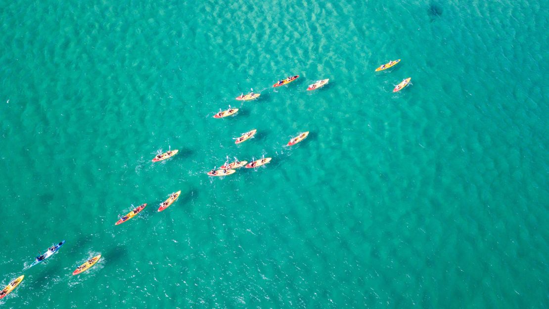 Aerial view of multiple colorful kayaks with paddlers rowing on clear turquoise water.