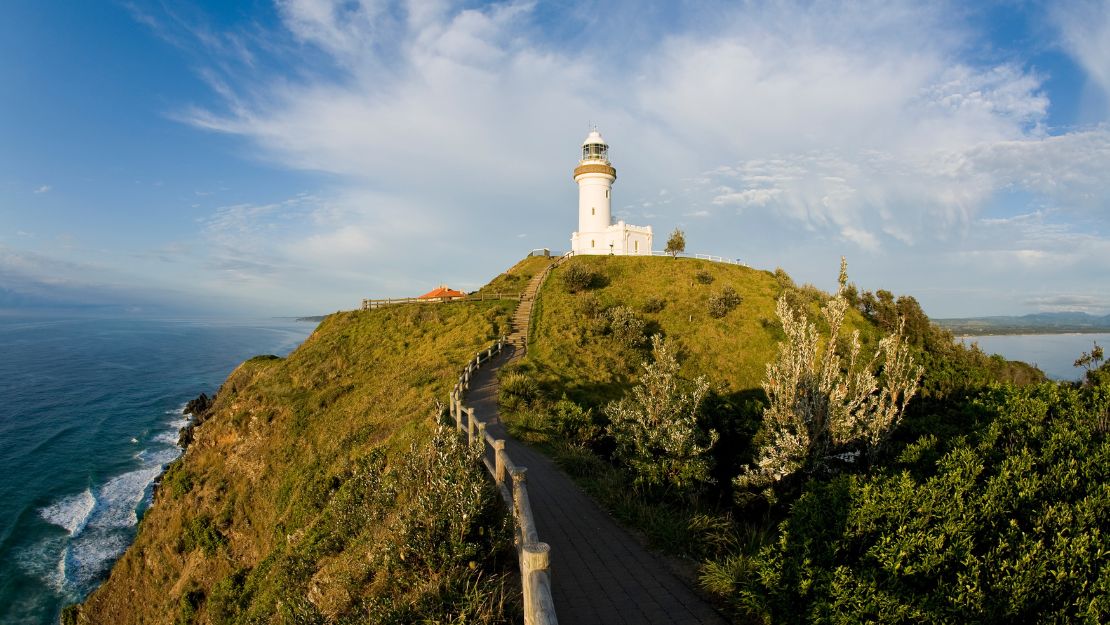 Lighthouse atop a green hill with a winding path, overlooking the ocean under a partly cloudy blue sky.