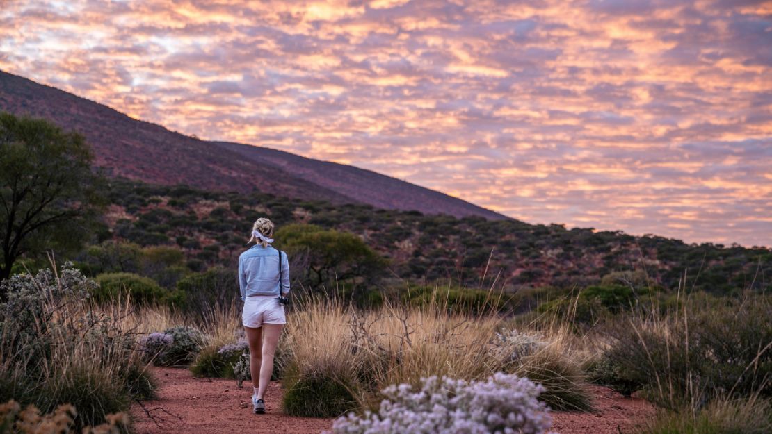Person walking on a trail through dry grass and shrubs with hills under a colorful, cloud-filled sunset sky.