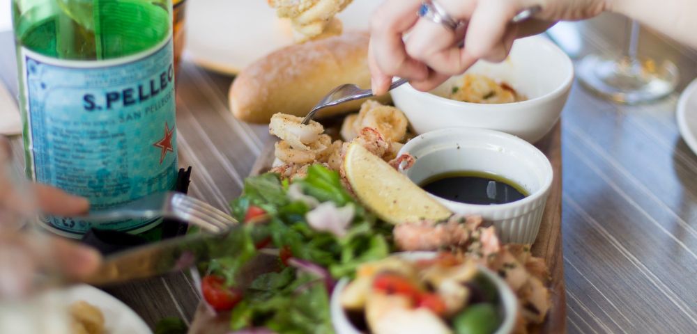 A table with a hand reaching for fried seafood near dipping sauce, fresh salad, lemon wedge, and a bottle of sparkling water, conveying a casual dining scene.