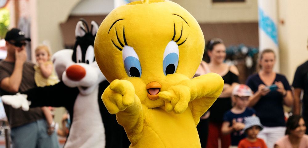 Bright yellow cartoon bird costume character, Tweety, playfully posing with hands up. Crowd of people smiling in background at an event. Cheerful atmosphere.