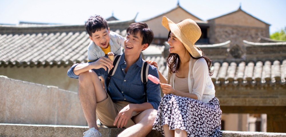 A happy family sits on stone steps enjoying ice cream. The father and son wear casual clothing; the mother, in a hat and skirt, smiles warmly. Background shows traditional architecture under a clear sky.