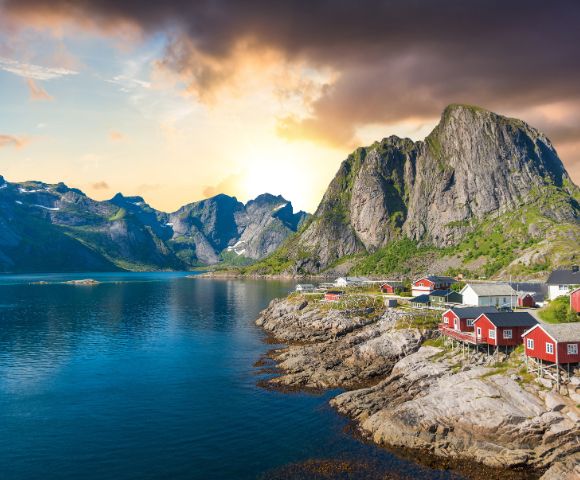 Coastal village at sunrise with red cabins on rocky shore, reflecting in the calm water. Majestic mountains in the background under a dramatic sky.