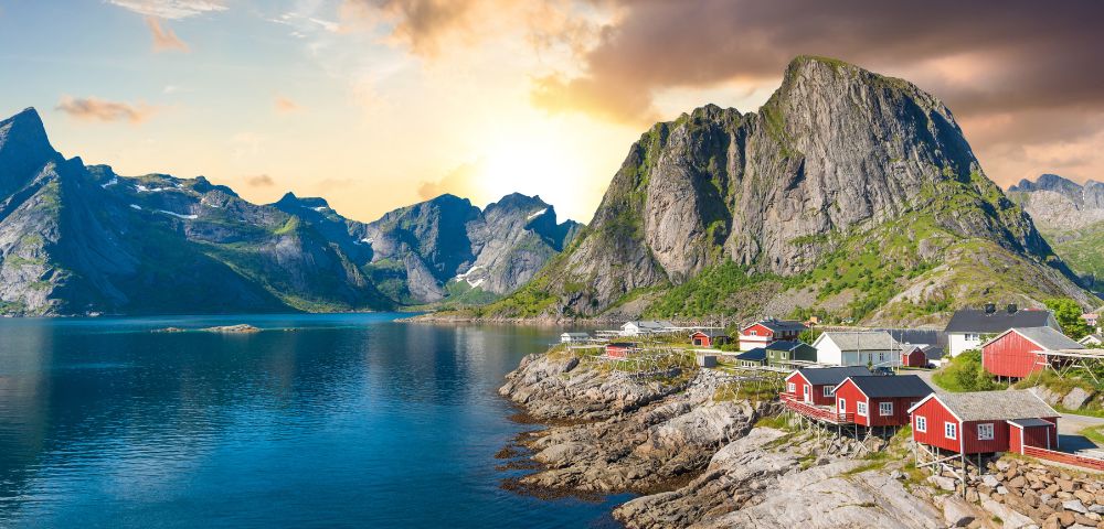 Coastal village at sunrise with red cabins on rocky shore, reflecting in the calm water. Majestic mountains in the background under a dramatic sky.