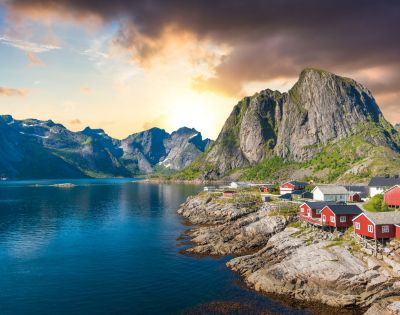 Coastal village at sunrise with red cabins on rocky shore, reflecting in the calm water. Majestic mountains in the background under a dramatic sky.