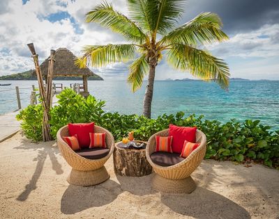 Tropical beach scene with two wicker chairs and a table under a palm tree, overlooking turquoise ocean. Chairs have red cushions, creating a relaxing vibe.
