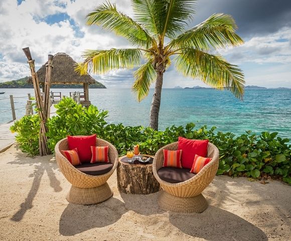 Tropical beach scene with two wicker chairs and a table under a palm tree, overlooking turquoise ocean. Chairs have red cushions, creating a relaxing vibe.
