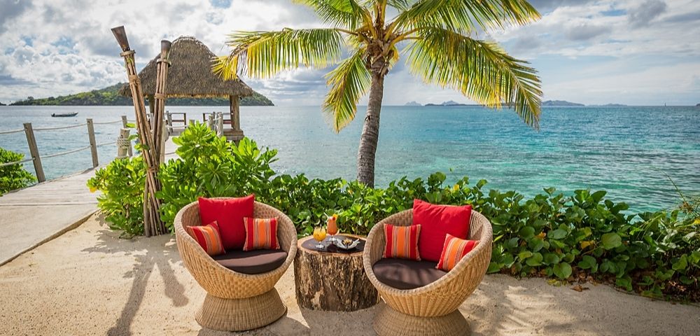 Tropical beach scene with two wicker chairs and a table under a palm tree, overlooking turquoise ocean. Chairs have red cushions, creating a relaxing vibe.