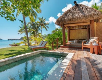 Tropical beachside scene with a private pool and two lounge chairs under palm trees. A thatched-roof cabana with seating overlooks turquoise water.