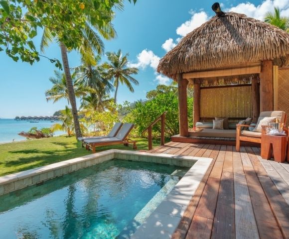 Tropical beachside scene with a private pool and two lounge chairs under palm trees. A thatched-roof cabana with seating overlooks turquoise water.