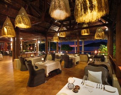 Tropical restaurant at sunset with wicker chairs and white tablecloths, mood lighting from woven pendant lights, and ocean view through open windows.