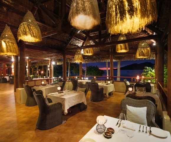 Tropical restaurant at sunset with wicker chairs and white tablecloths, mood lighting from woven pendant lights, and ocean view through open windows.
