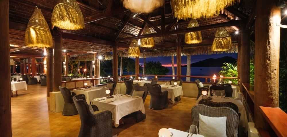 Tropical restaurant at sunset with wicker chairs and white tablecloths, mood lighting from woven pendant lights, and ocean view through open windows.