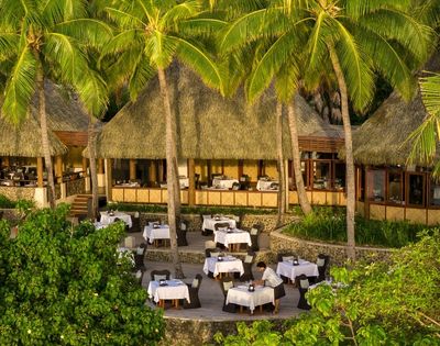 A tropical outdoor restaurant with white tablecloth-covered tables under thatched roofs. Surrounded by lush greenery and palm trees, it has a serene ambiance.