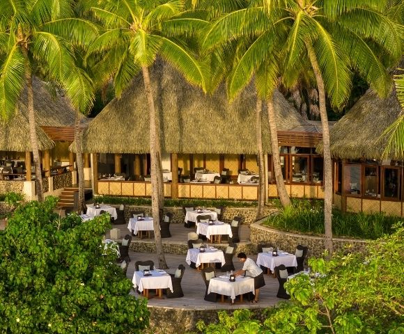 A tropical outdoor restaurant with white tablecloth-covered tables under thatched roofs. Surrounded by lush greenery and palm trees, it has a serene ambiance.