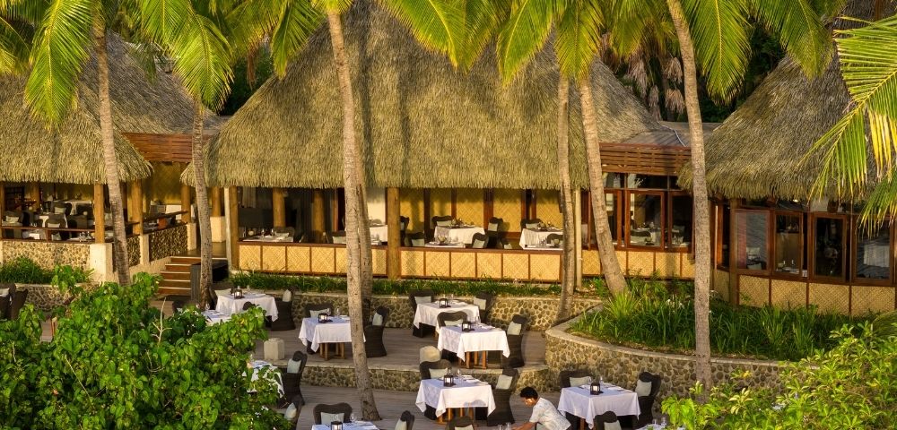 A tropical outdoor restaurant with white tablecloth-covered tables under thatched roofs. Surrounded by lush greenery and palm trees, it has a serene ambiance.