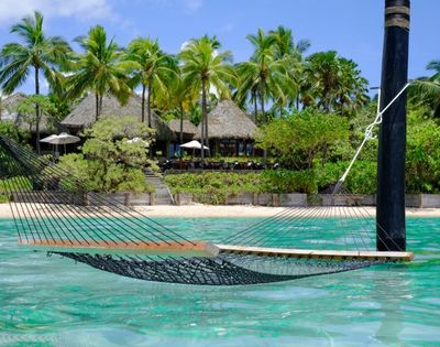 Hammock floating above turquoise water near a sandy beach, surrounded by lush green palm trees and a thatched-roof beachfront resort. Peaceful and tropical.