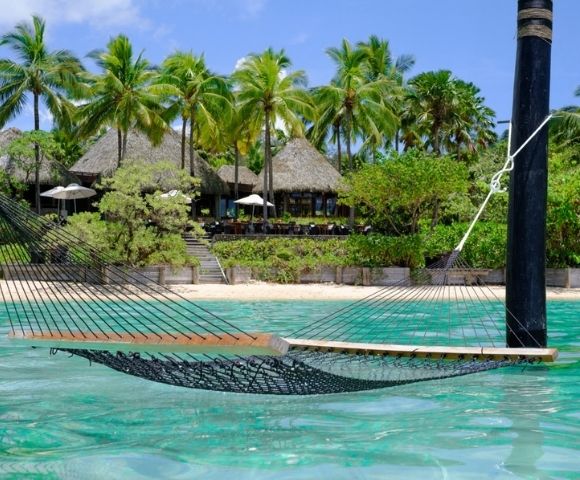 Hammock floating above turquoise water near a sandy beach, surrounded by lush green palm trees and a thatched-roof beachfront resort. Peaceful and tropical.